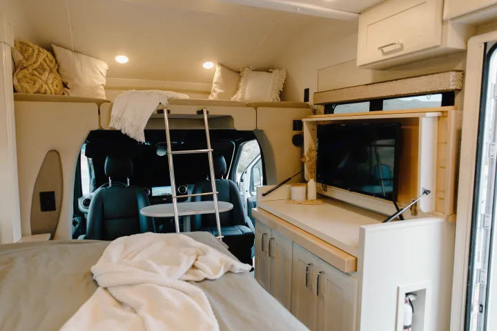Modern RV rental interior: large bed, upper bunk with ladder, compact kitchen counter with TV, and front vehicle seats.. Tiffin Motorhomes Wayfarer 25 JW 2021