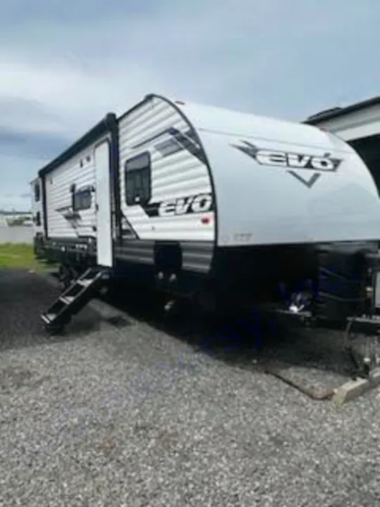 A modern white and grey EVO travel trailer, perfect for RV rental, is parked on a gravel lot. Steps are visible at the entrance.. Forest River Evo 2023