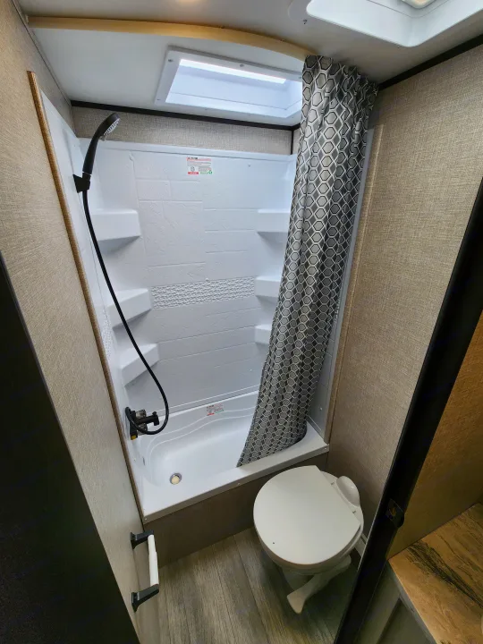 Interior of an RV rental bathroom featuring a shower stall with a patterned curtain, a toilet, and wood-look flooring.. Jayco Jay Flight 2023