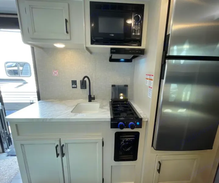 Compact RV rental kitchen showcasing a sink, two-burner stovetop, microwave, and refrigerator with white cabinets and backsplash.. Jayco Jay Flight 2023
