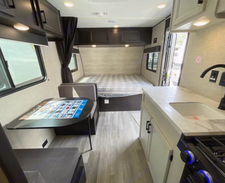 RV rental interior: A bed, dinette, and kitchen with sink/stove visible. Light wood-look flooring, dark cabinets.. Jayco Jay Flight 2023