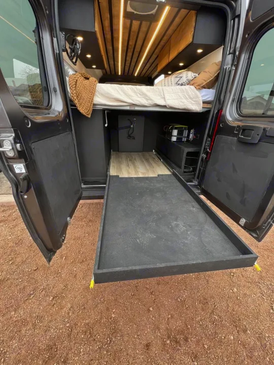Inside an RV rental: a cozy bed above a pull-out storage tray. Wood accents & lighting create a modern mobile. Dodge Ram Promaster 2500 2022