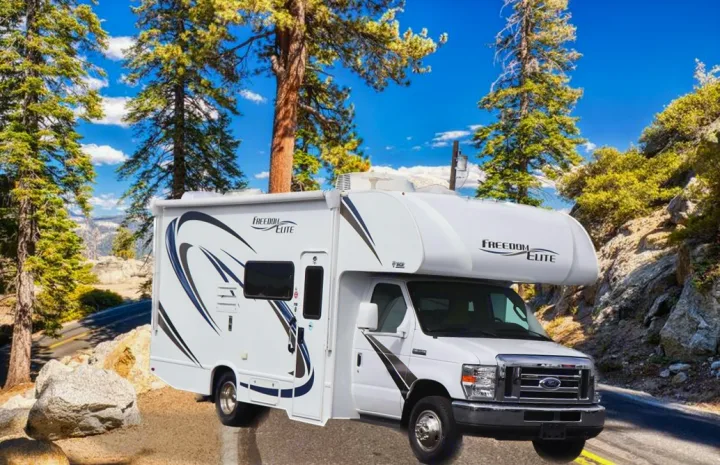 Experience nature with an RV rental! A white Freedom Elite RV on a scenic mountain road, surrounded by tall trees and rocks.. Thor Motor Coach Freedom Elite 2018