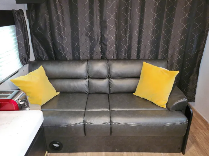 An RV rental's living area: a comfortable dark grey sofa with two bright yellow pillows and patterned curtains.. Forest River Cherokee Grey Wolf 2022