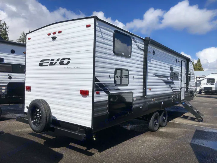 This white and grey EVO travel trailer, an ideal RV rental, is seen in a paved lot with a rear spare tire.. Forest River Evo 2023