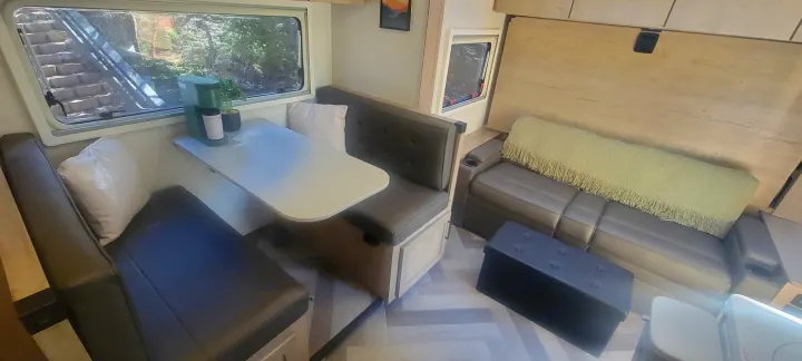 Interior of a bright rv rental featuring a dinette, sofa, and a scenic window looking out onto nature. Modern and cozy design.. Ember Overland 2023