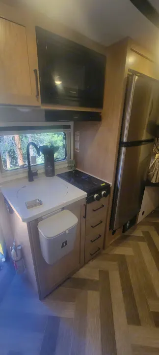 Inside an RV rental: a compact kitchen with a sink, microwave, 2-burner stove, and fridge in light wood cabinetry.. Ember Overland 2023