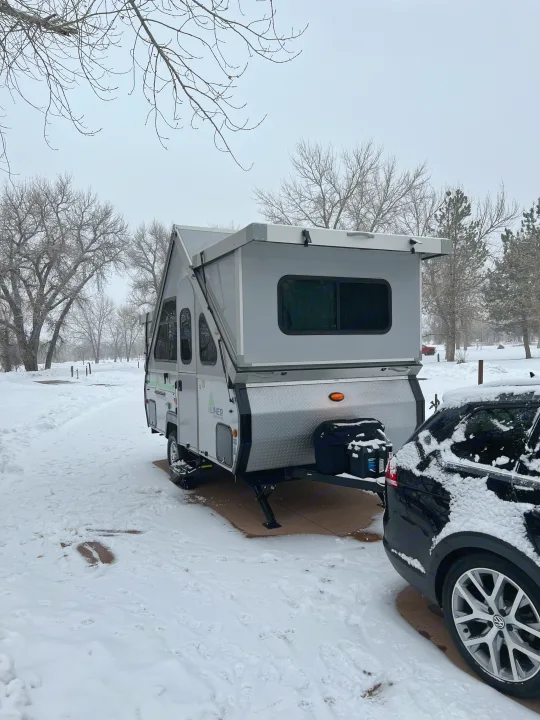 Winter camping: An A-frame RV rental is parked in a snowy campground next to a snow-covered car.. A-liner Classic 2022