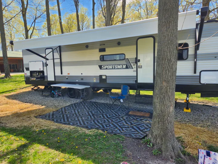 Spacious RV rental on a gravel campsite with extended awning, outdoor mat, table, and chair. Ideal for a relaxing outdoor trip.. Sportsmen Other 2022
