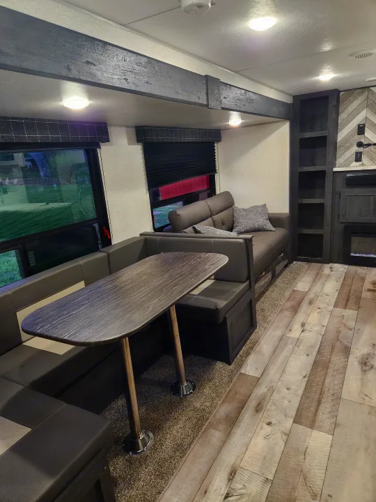 Modern RV rental living space: comfy dinette, sofa, wood accents, and bright lighting. Large window shows green outdoors.. Sportsmen Other 2022
