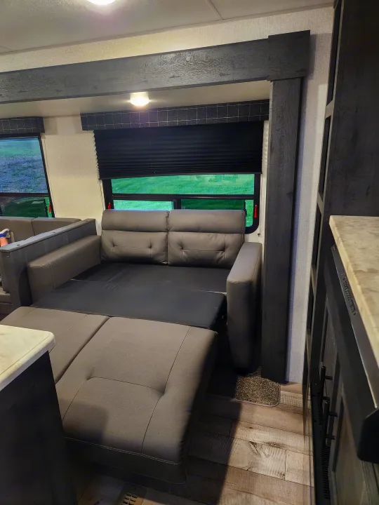 Spacious RV rental interior featuring a modern gray sofa/bed, large windows with green views, and wood-look flooring.. Sportsmen Other 2022