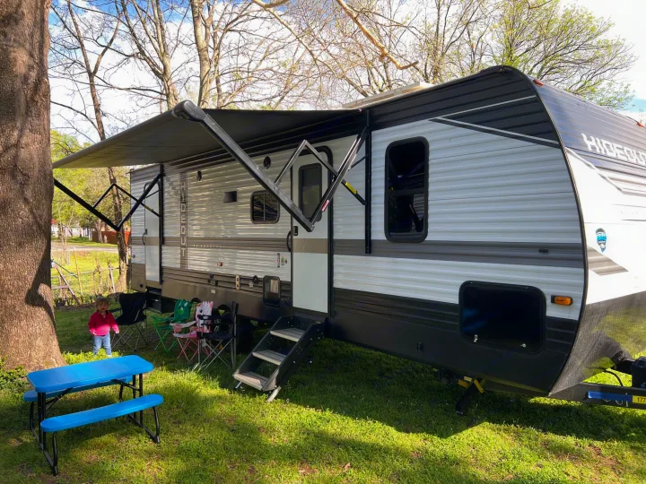 Outdoor adventure with an RV rental. A modern travel trailer with awning deployed, a child, and picnic table at a grassy campsite.. Keystone Hideout 2022