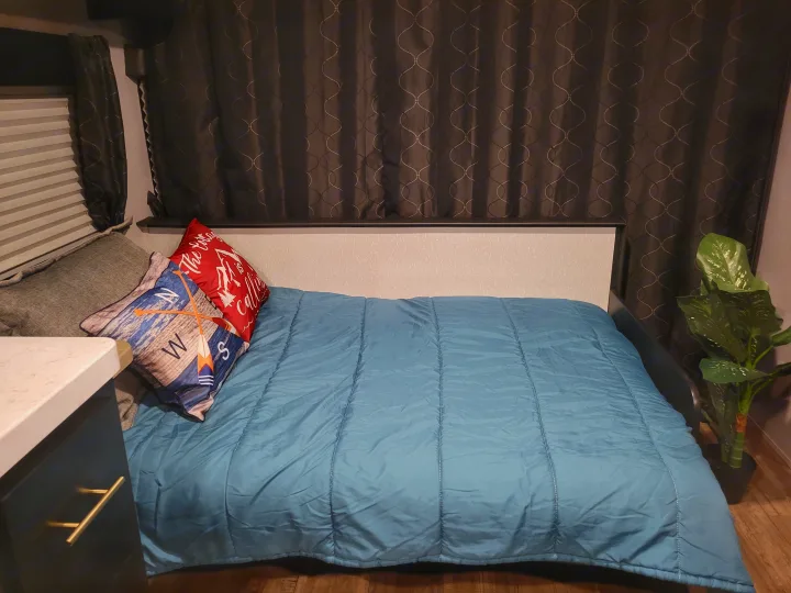 Cozy RV rental bedroom featuring a blue comforter, patterned pillows, dark curtains, and a faux plant.. Forest River Cherokee Grey Wolf 2022