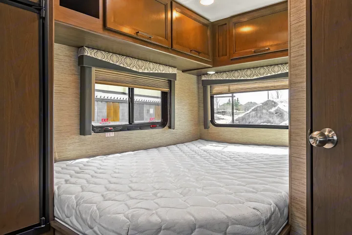 Inside a cozy RV rental bedroom: quilted white mattress, wooden cabinets, and windows offering a winter scene outside.. Thor Freedom Elite 2021