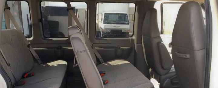 Inside a spacious RV rental van, showing rows of grey passenger seats. Outside, a white truck and industrial area are visible.. Chevrolet Express 1500 2009
