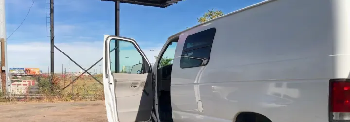 White van, possibly an RV rental, with its door open near a large billboard structure under a clear blue sky.. Ford E-150 2000