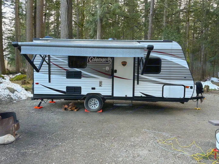White Coleman travel trailer (RV) with awning extended, parked in a snowy forest campground. Ideal rv rental for your camping trips!. Dutchmen Coleman Lantern LT 2019