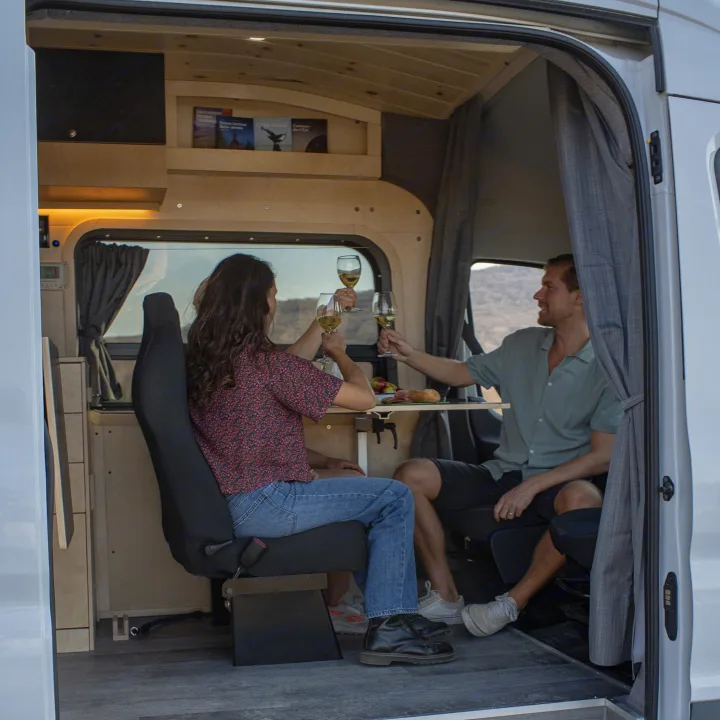 Couple enjoying wine and food in a cozy RV rental. This camper van offers a comfortable dining experience for travelers.. Ford Van 2023