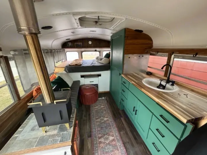 Converted bus RV rental showcasing a modern kitchen with teal cabinets, wood stove, and rear bed setup.. GMC Savana 2002