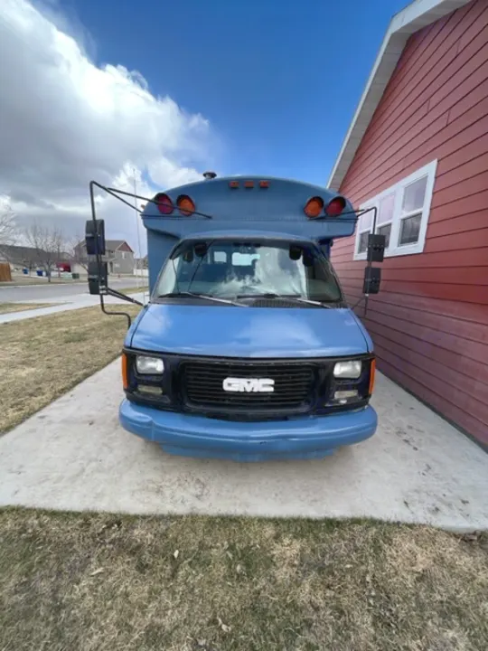 A blue GMC short bus, ideal for an RV rental, parked by a red house on a sunny day with scattered clouds.. GMC Savana 2002