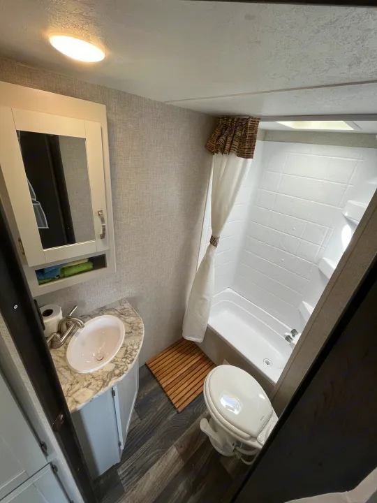 Bright RV rental bathroom with white fixtures: sink, toilet, and tub/shower. Features light grey walls and dark wood floor.. Keystone RV Bullet Ultra Lite 2019