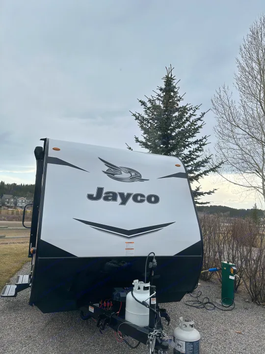 A white Jayco travel trailer (RV rental) parked on gravel with propane tanks and an electrical hookup visible.. Jayco Jay Flight SLX 2022