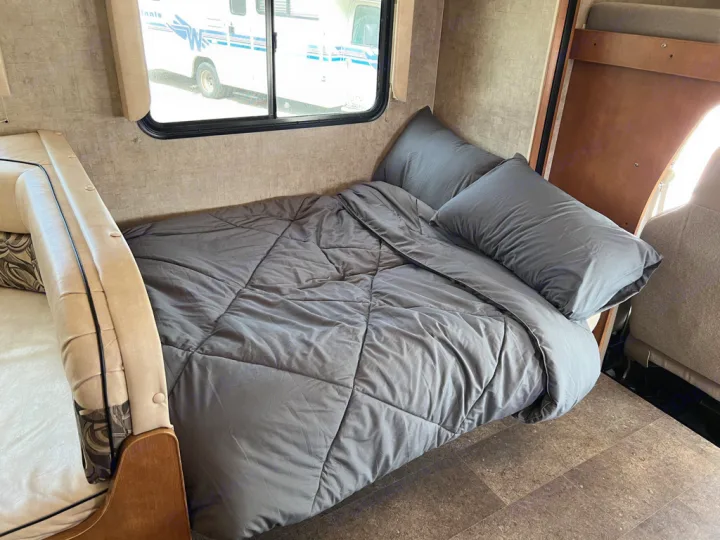 Comfortable sleeping area inside an RV rental, featuring a cozy bed with grey comforter and pillows, beside a window view.. Winnebago Minnie Winnie 2015