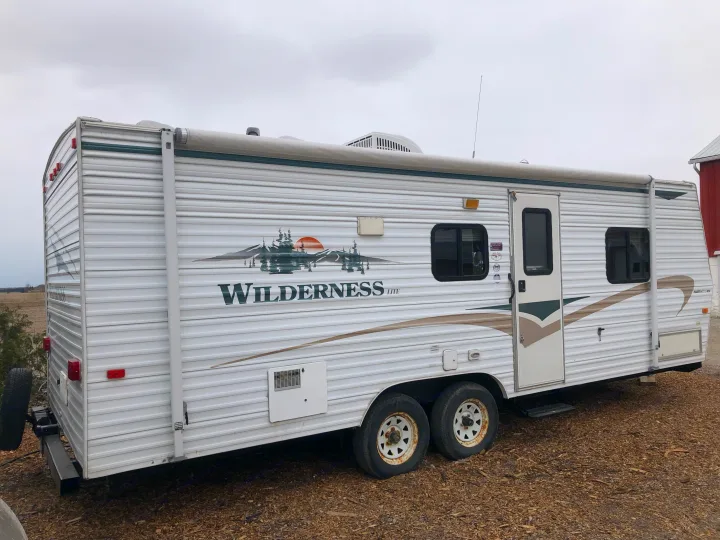 White Wilderness travel trailer for rv rental, with rolled awning, parked outdoors on wood chips. Rear spare tire visible.. Wilderness Lite 2003