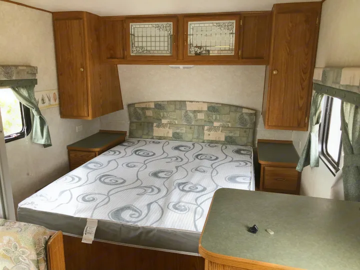 This RV rental bedroom features a patterned mattress, wooden cabinets, and green curtains by the window.. Wilderness Lite 2003