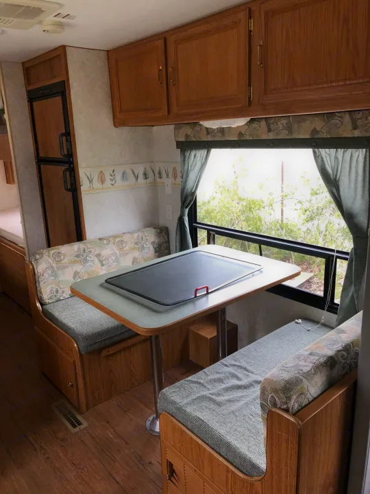 Inside an RV rental, featuring a dinette with patterned benches, wooden cabinets, a window view of trees, and a refrigerator.. Wilderness Lite 2003