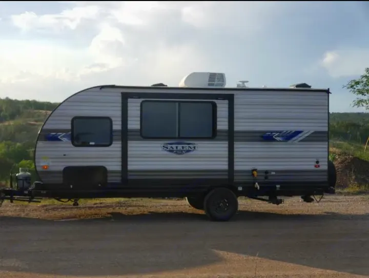 Side view of a modern gray Forest River Salem travel trailer RV rental on a gravel road, amidst natural scenery.. Forest River Salem FSX 178bhsk 2023
