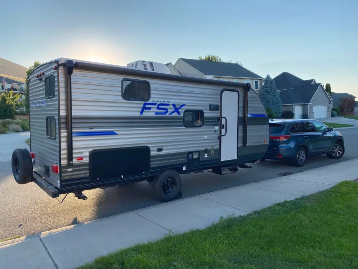 Grey Salem FSX travel trailer (RV rental) hitched to a blue SUV on a residential street, with houses and hills behind.. Forest River Salem FSX 178bhsk 2023