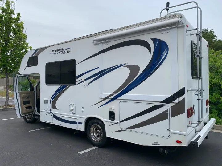 White RV rental with blue/gray graphics parked on asphalt, driver's door open, showing side features like an awning and rear ladder.. Thor Freedom Elite 2019