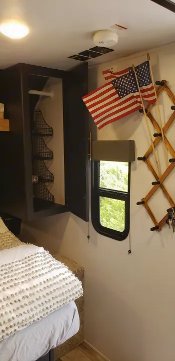 Cozy RV rental bedroom with a bed, open overhead cabinet storage, window, American flags, and a key rack.. Keystone RV Springdale 2022
