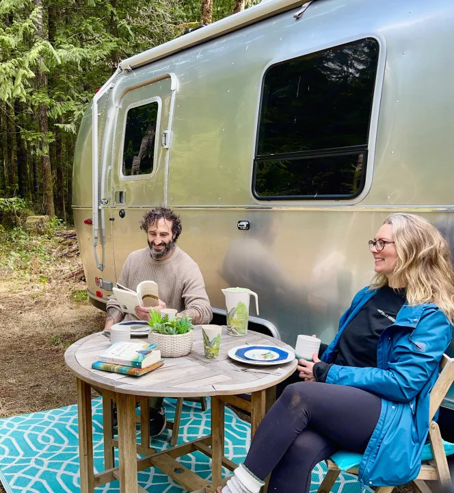 Couple enjoys reading and coffee at a table by their Airstream RV rental, nestled in a serene forest. Ideal for glamping.. Airstream Sport 2019