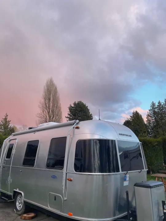 A shiny Airstream travel trailer (RV rental) under a vibrant pink and blue sunset sky, surrounded by tall trees.. Airstream Sport 2019