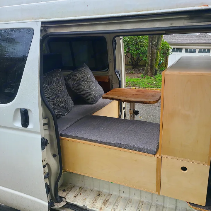 A vertical view of an RV rental camper van's interior. Features custom wood cabinets, grey seating, and a table.. Toyota Hiace 2006