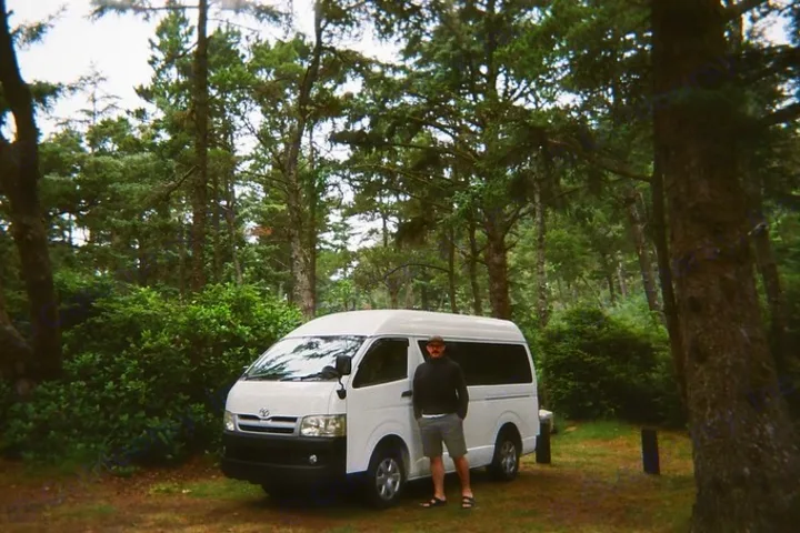 Man stands by a white RV rental van in a lush forest campsite. Perfect for outdoor adventure and travel.. Toyota Hiace 2006