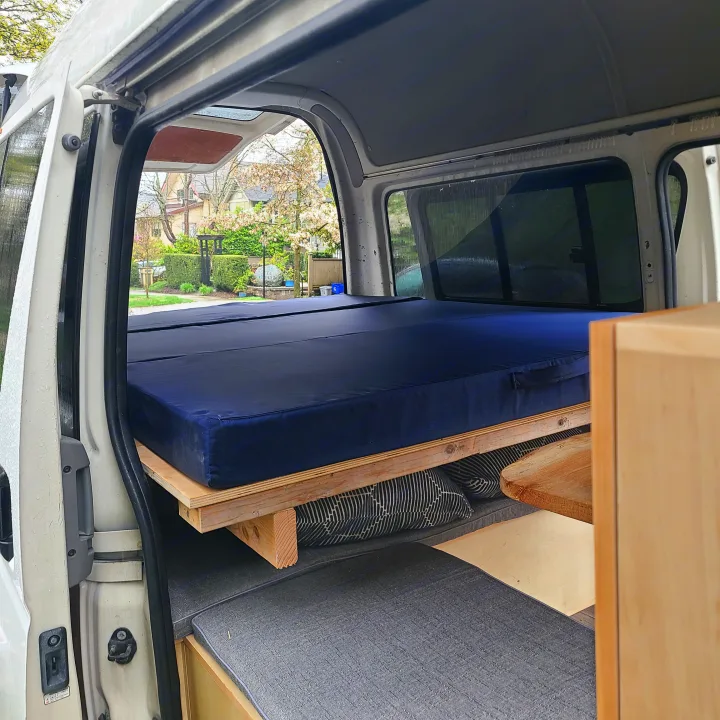 Sideways view inside an rv rental camper van: A blue bed, wooden frame, and grey cushions. Open doors show an outdoor scene.. Toyota Hiace 2006