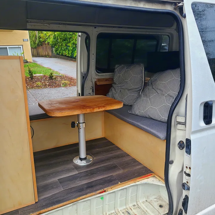 rv rental: Inside a custom camper van, featuring wooden benches, a dark laminate floor, patterned cushions, and a removable table.. Toyota Hiace 2006