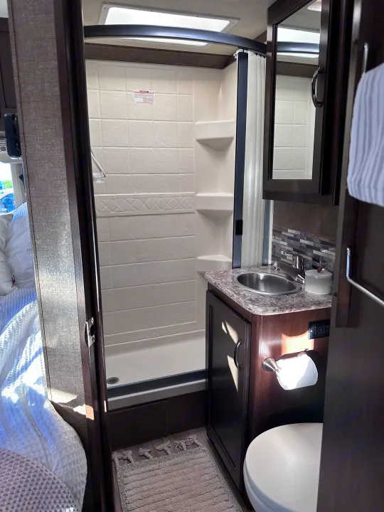 Inside an RV rental, this clean, compact bathroom features a tiled shower, sink with mirror, and toilet.. Thor Four Winds 2020
