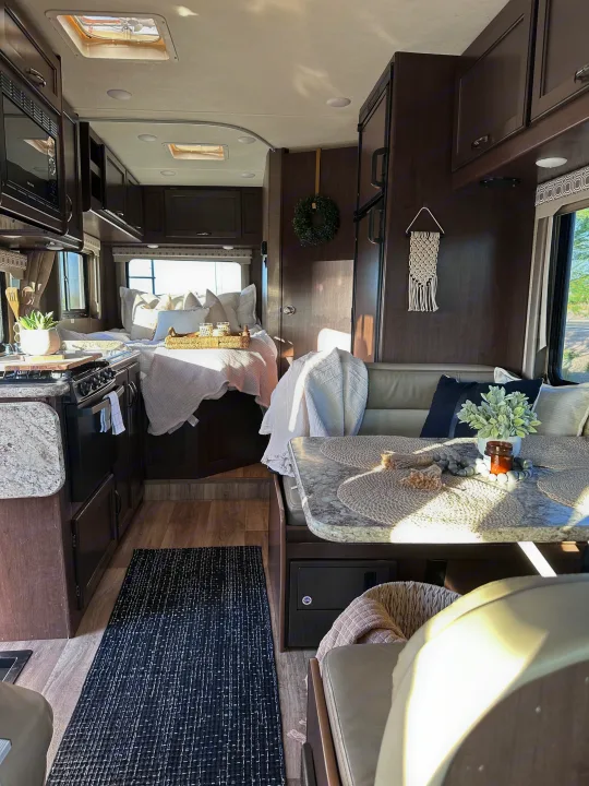 Bright RV rental interior with a compact kitchen, bed with white bedding, and dining area. Features dark wood and a patterned rug.. Thor Four Winds 2020