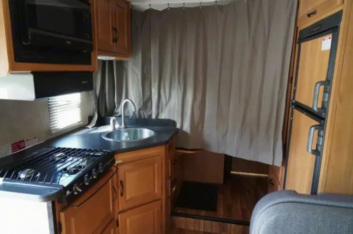 Inside an RV rental, a compact kitchen with a stove, sink, microwave, and fridge, separated by a light privacy curtain.. Thor Majestic 2012