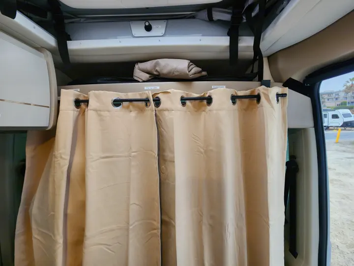Inside an RV rental, featuring beige privacy curtains, overhead storage labeled 'Pop-top Ladder' & 'Window Covers'.. Thor Tellaro 2023