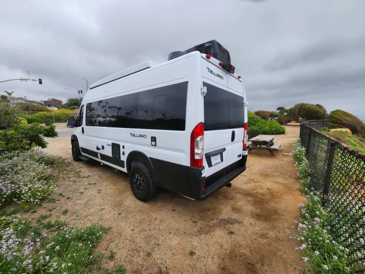 This white Tellaro RV rental is parked on a dirt lot by a picnic table and a fence amidst green plants under a cloudy sky.. Thor Tellaro 2023