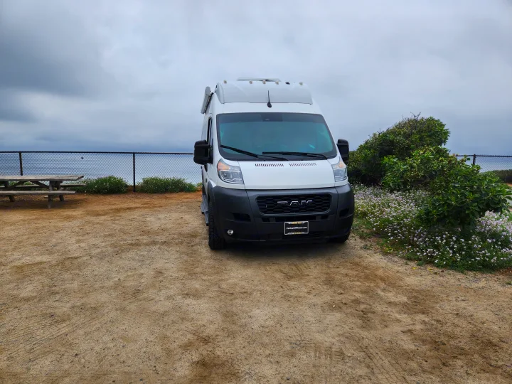 White camper van RV rental parked on a coastal dirt lot with ocean views, a fence, and picnic table.. Thor Tellaro 2023