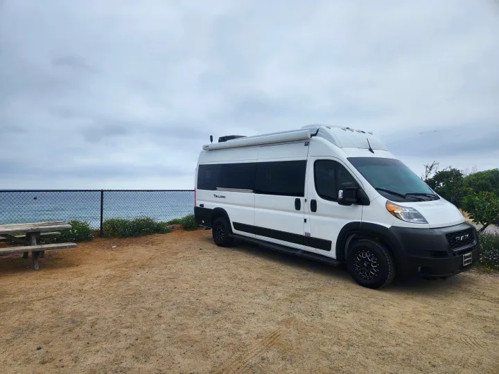 White **RV rental** van parked on a dirt lot overlooking the ocean, with a picnic table and cloudy sky.. Thor Tellaro 2023