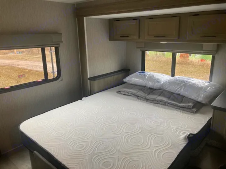 Cozy RV rental bedroom with a new mattress, pillows, and blanket on the bed. Windows look out to the outdoors.. Coachmen 26DS 2021