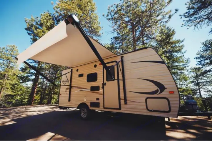 This cream-colored RV rental, a travel trailer with an extended awning, is parked outdoors amidst tall pine trees.. Keystone RV Hideout 2018