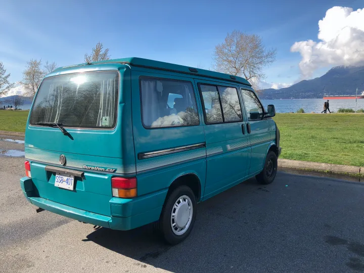 This teal Volkswagen EuroVan CV camper van RV rental is parked in a scenic park by the water with distant mountains.. Volkswagen T4 Westfalia Coach 1992